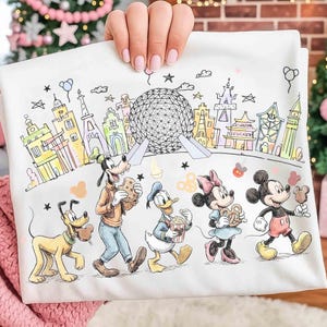May include: White t-shirt featuring a colorful illustration of Disney characters and landmarks. Pluto, Goofy, Donald Duck, Minnie Mouse, and Mickey Mouse are depicted with food items. The background includes the Eiffel Tower and Spaceship Earth.