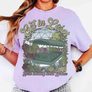 Könnte beinhalten: Helllila T-Shirt mit einem Grafikdesign einer Bootsfahrt, mit dem Text "Get in Lose" und "The Land". Auf dem unteren Teil des Shirts steht "We're learning about Lettuce". Das Design enthält grüne, braune und violette Elemente.