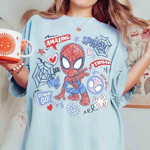 May include: Light blue t-shirt featuring a cartoon Spider-Man design. The graphic includes the word "AMAZING" in a red banner, spiderwebs, hearts, and the words "SPIDER" and "GO-WEBS-GO!"