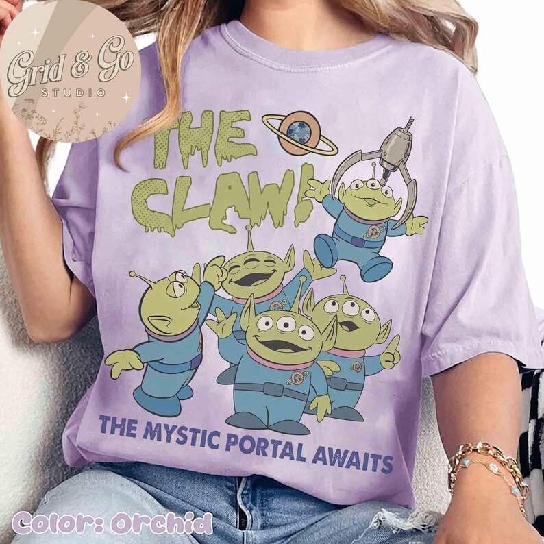 May include: Orchid-colored t-shirt featuring a graphic of the aliens from the movie Toy Story. The text "THE CLAW!" and "THE MYSTIC PORTAL AWAITS" are printed on the shirt. The aliens are green with blue uniforms.