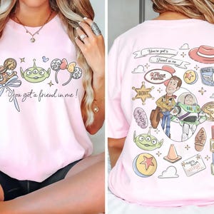 Op de afbeelding: Roze T-shirt met een afbeelding van stripfiguren op de voorkant en de tekst "You got a friend in me!". De achterkant van het shirt heeft meer stripfiguren en tekst.