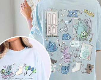May include: Light blue t-shirt featuring a colorful cartoon graphic. The design includes a door, monsters, and the text "We scare because we care!" The shirt has a crew neck and short sleeves.
