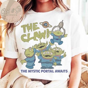 May include: White t-shirt featuring a cartoon graphic of green aliens in blue suits with the text "THE CLAW!" and "THE MYSTIC PORTAL AWAITS". The aliens have three eyes and are reaching upwards.