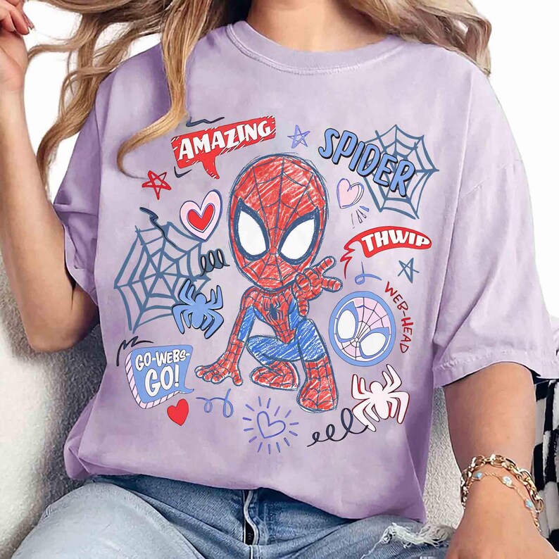 May include: A light purple t-shirt featuring a cartoon Spider-Man design. The graphic includes the word "AMAZING" in a red speech bubble, spiderwebs, hearts, and the word "SPIDER". The shirt has short sleeves.