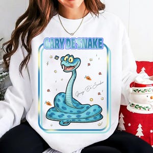 Puede incluir: Sudadera blanca con un dise&ntilde;o de serpiente de dibujos animados. La serpiente azul con lunares azules oscuros tiene una cara sonriente, con el texto "GARY DE SNAKE" encima. Peque&ntilde;as estrellas y zanahorias rodean la serpiente.