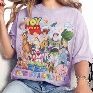 May include: A lilac t-shirt with a vibrant graphic of characters from the animated film "Toy Story." The design includes Woody, Buzz Lightyear, and other characters, with the text "TOY STORY" and "YOU'VE GOT A FRIEND IN ME."