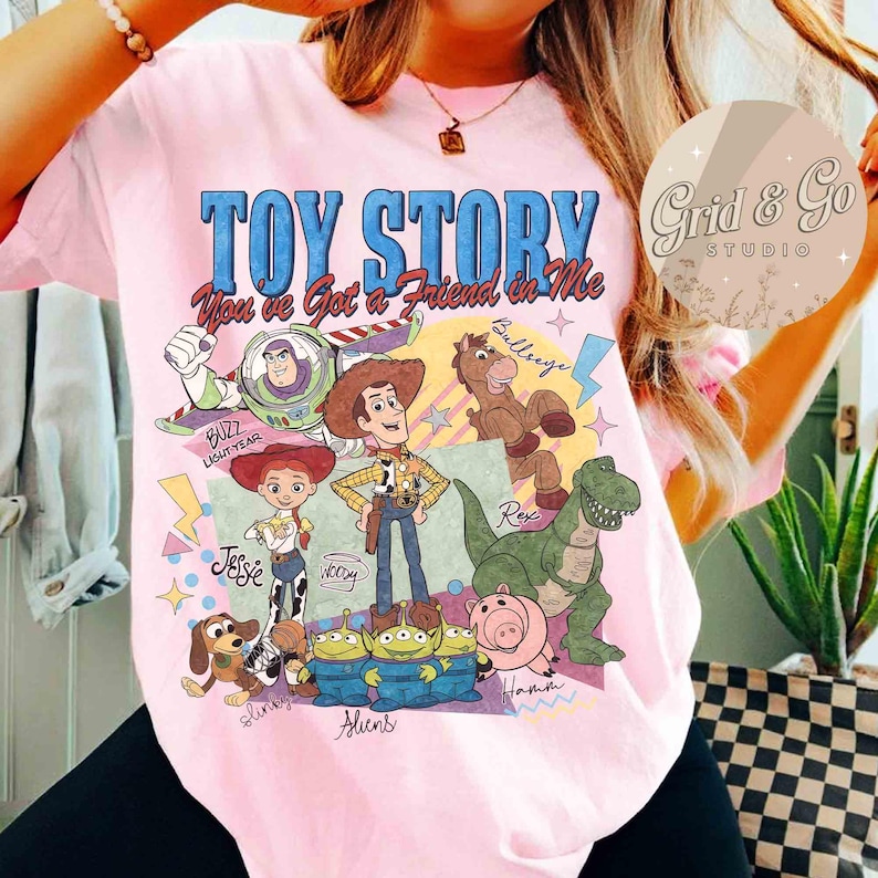 May include: Pink t-shirt featuring a graphic of Toy Story characters, including Woody, Buzz Lightyear, and Rex. The text "Toy Story You've Got a Friend in Me" is displayed in blue. The shirt has a crew neck and short sleeves.