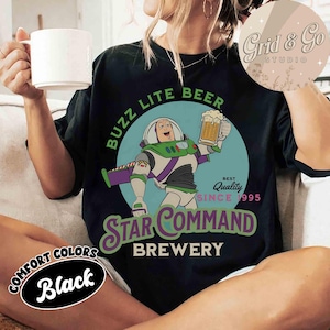 以下が含まれることがあります： 黒いTシャツ。宇宙レンジャーの漫画がビールジョッキを持っています。Tシャツには「BUZZ LITE BEER STAR COMMAND BREWERY」と書かれており、「BEST QUALITY SINCE 1995」と「COMFORT COLORS Black」がプリントされています。