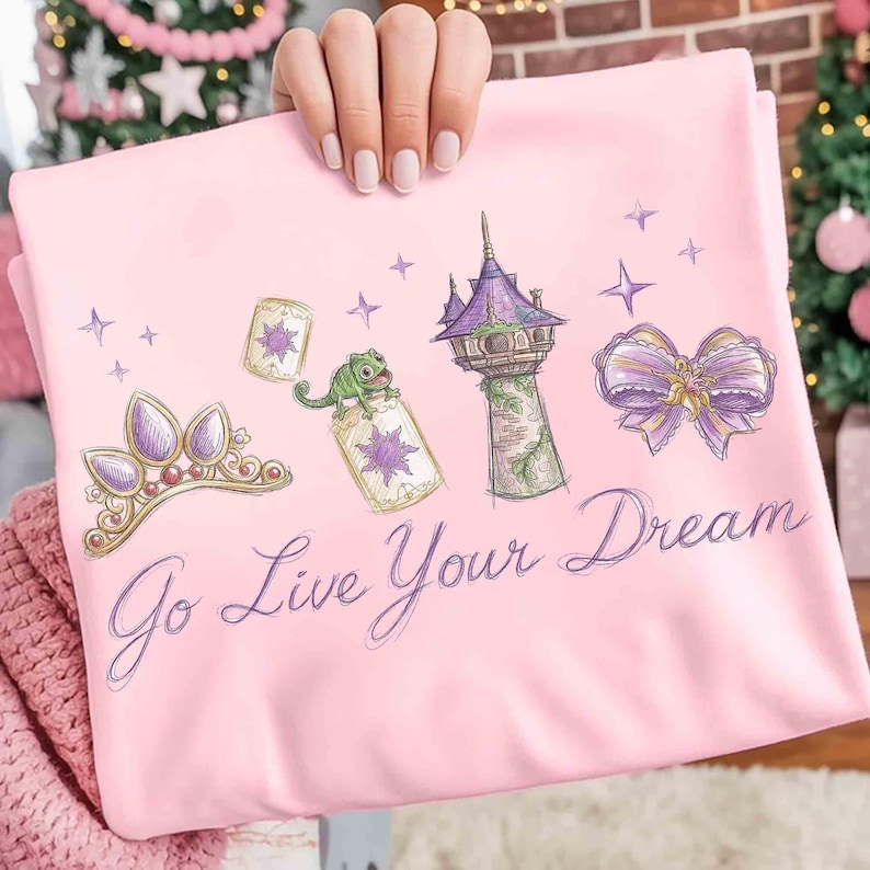 Może przedstawiać: Jasnor&oacute;żowa koszulka z kresk&oacute;wkowym wzorem przedstawiającym fioletową tiarę, wieżę, kokardę i kameleona. Tekst "Go Live Your Dream" jest napisany kursywą. Projekt jest w stylu kresk&oacute;wkowym.