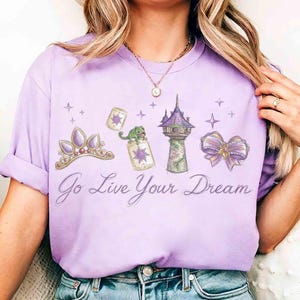 Może przedstawiać: Jasnofioletowa koszulka z grafiką inspirowaną bajką. Projekt zawiera tiarę, żabę, wieżę, kokardę oraz napis "Go Live Your Dream" w ozdobnej czcionce. Koszulka jest w stylu casual.