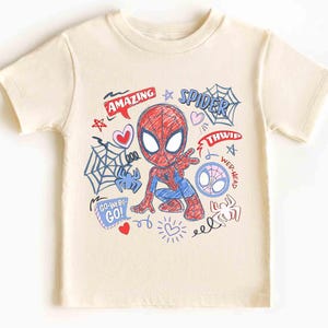 May include: Cream-colored t-shirt featuring a colorful graphic of Spider-Man in a crouching pose. The design includes the words "AMAZING" and "SPIDER", along with spiderwebs, hearts, and other doodles. The shirt is short-sleeved.