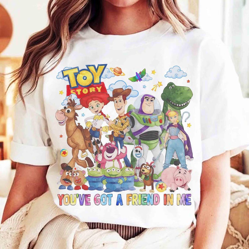 May include: White t-shirt with a vibrant graphic of "Toy Story" characters, including Woody and Buzz Lightyear. The design features the film's title and the phrase "YOU'VE GOT A FRIEND IN ME" in colourful lettering.