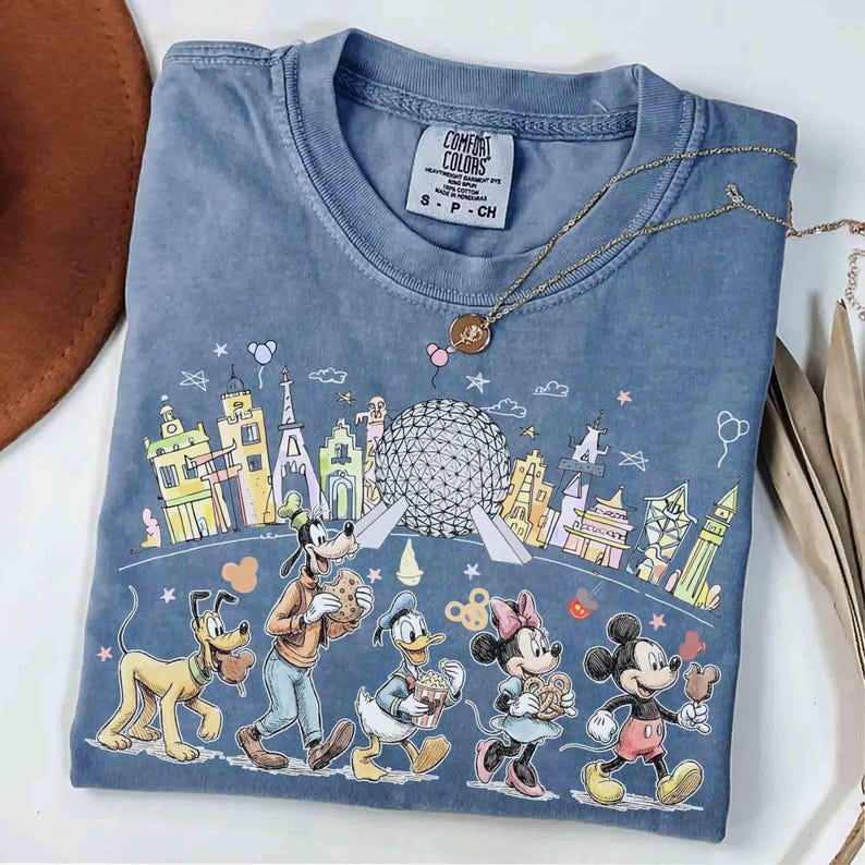 May include: A blue Comfort Colors t-shirt featuring a colorful graphic of Disney characters walking in front of a cityscape. Pluto, Goofy, Donald Duck, Minnie Mouse, and Mickey Mouse are depicted. A gold necklace with a pendant is draped on the shirt.