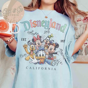 Puede incluir: Camiseta azul claro con la palabra "Disneyland" en colores pastel, junto con los personajes de dibujos animados Mickey Mouse, Minnie Mouse, Goofy, Pluto y Donald Duck. La camiseta tambi&eacute;n incluye el texto "EST. 1959" y "CALIFORNIA".