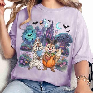 May include: A light purple t-shirt featuring a graphic print of two chipmunks in Halloween costumes, one as a mummy and one as a pumpkin, standing before a theme park castle, a geodesic sphere, and a spooky tree with bats and balloons.