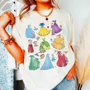 以下が含まれることがあります： クリーム色のTシャツ。様々なプリンセスドレスのカラフルなイラストが特徴です。デザインには、青、黄、緑、赤のドレスと、キャラクターの詳細が含まれています。このシャツはカジュアルで快適なスタイルです。