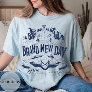 Op de afbeelding: Een lichtblauw T-shirt met korte mouwen en een donkerblauwe grafische print van de Hulk en Spider-Man met een stadsskyline op de achtergrond. De tekst "BRAND NEW DAY" staat in grote donkerblauwe hoofdletters in het midden.