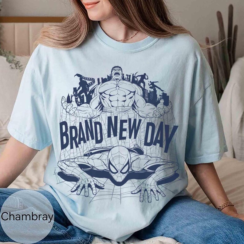 Op de afbeelding: Een lichtblauw T-shirt met korte mouwen en een donkerblauwe grafische print van de Hulk en Spider-Man met een stadsskyline op de achtergrond. De tekst "BRAND NEW DAY" staat in grote donkerblauwe hoofdletters in het midden.