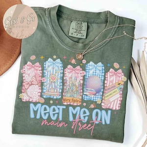Pode incluir: Camiseta verde Comfort Colors com um gráfico com ícones de parques temáticos e o texto "Meet Me on Main Street". O design inclui uma roda gigante, um castelo e cartões de embarque. Um colar de prata com um pingente está na camisa.