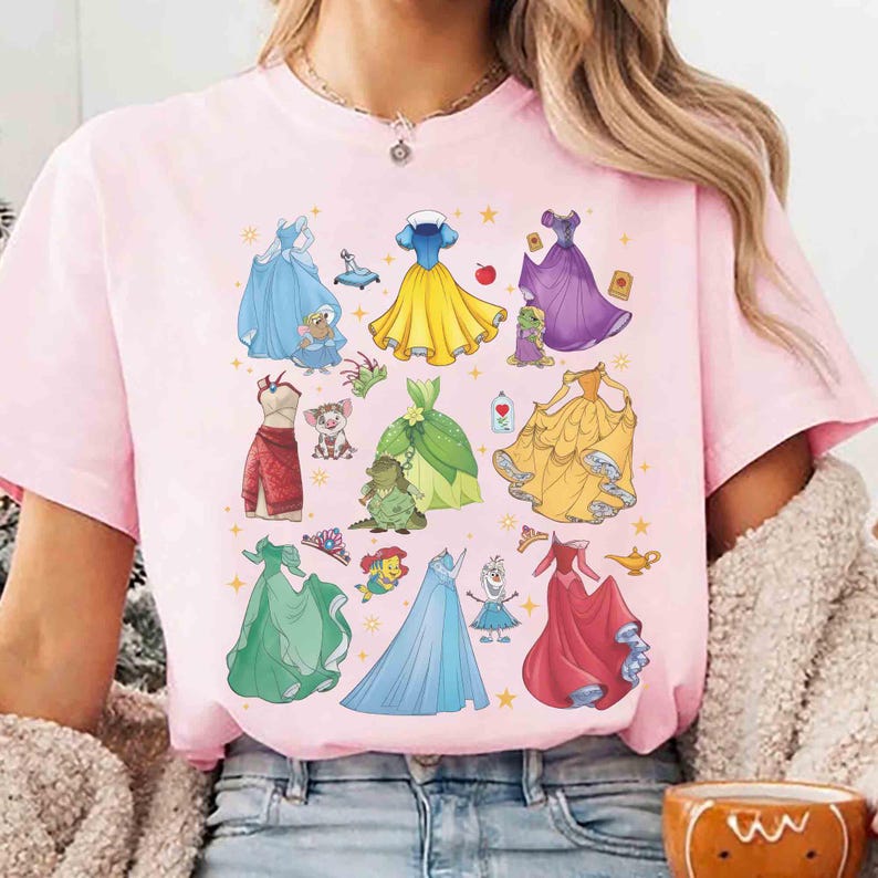 以下が含まれることがあります： 様々なプリンセスドレスと関連アイコンのカラフルなグラフィックが特徴のピンクのTシャツ。デザインには、青、黄、緑、赤のドレスと、キャラクターに関連する画像が含まれています。カジュアルウェアアイテムです。
