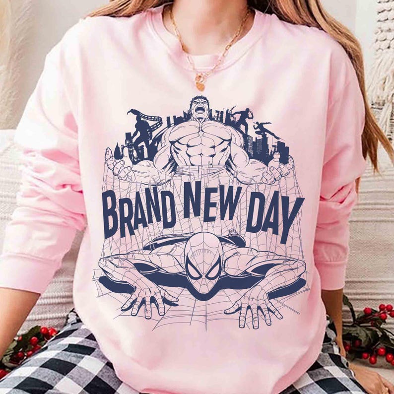 Op de afbeelding: Een lichtroze sweater met ronde hals en een donkerblauwe grafische print van de Hulk en Spider-Man. De tekst "BRAND NEW DAY" staat in het midden gedrukt. Het ontwerp bevat een spinnenwebpatroon en silhouetten van stadsgebouwen.