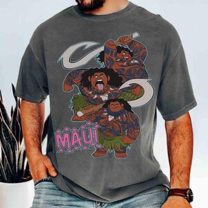 May include: Gray t-shirt featuring a cartoon illustration of Maui from the movie Moana. The design includes three figures of Maui with blue tribal tattoos, a fish hook, and the word "MAUI" in pink letters.