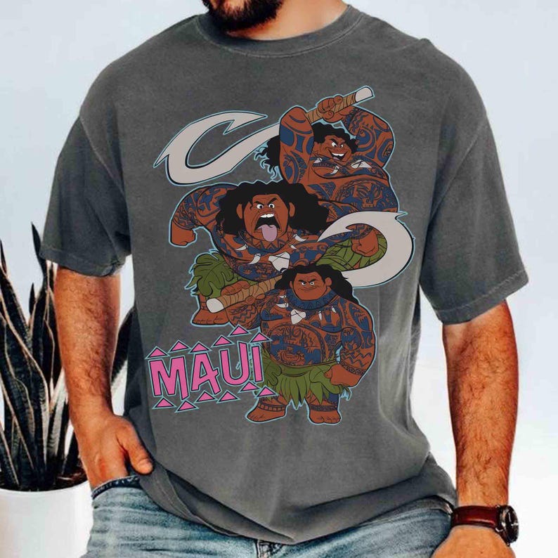 May include: Gray t-shirt featuring a cartoon illustration of Maui from the movie Moana. The design includes three figures of Maui with blue tribal tattoos, a fish hook, and the word "MAUI" in pink letters.