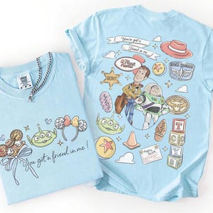 Op de afbeelding: Twee lichtblauwe t-shirts met cartoonafbeeldingen. E&eacute;n heeft personages en iconen uit een film, de andere heeft een strik, een alien en de tekst "You got a friend in me!". Een zilveren kettingketting ligt op &eacute;&eacute;n shirt.