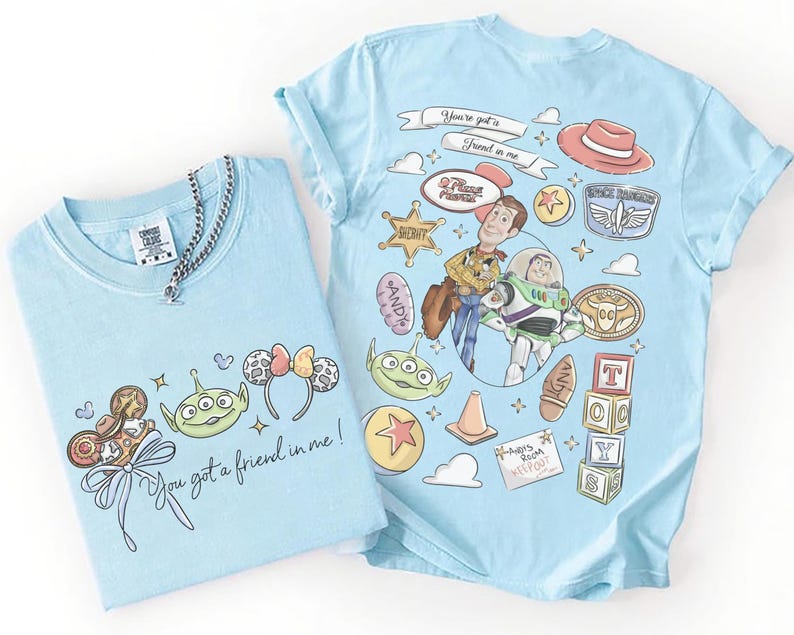 Op de afbeelding: Twee lichtblauwe t-shirts met cartoonafbeeldingen. E&eacute;n heeft personages en iconen uit een film, de andere heeft een strik, een alien en de tekst "You got a friend in me!". Een zilveren kettingketting ligt op &eacute;&eacute;n shirt.