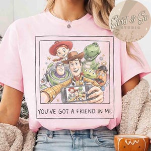 May include: Pink t-shirt featuring a graphic of Toy Story characters taking a selfie. The image includes Woody, Buzz Lightyear, Jessie, Rex, and other characters. The text "YOU'VE GOT A FRIEND IN ME" is printed below the image.