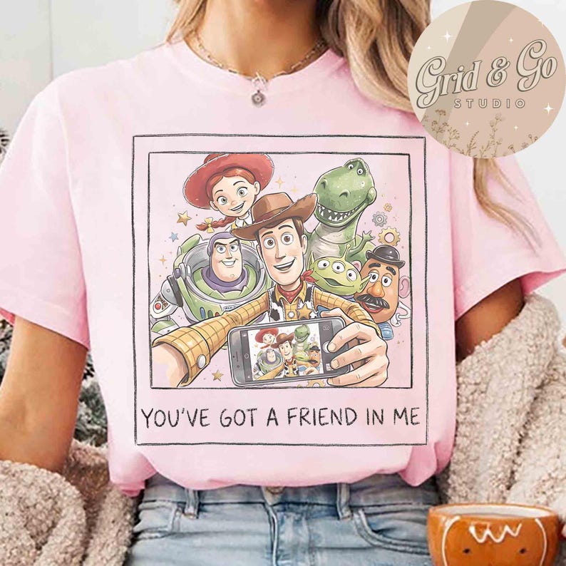 May include: Pink t-shirt featuring a graphic of Toy Story characters taking a selfie. The image includes Woody, Buzz Lightyear, Jessie, Rex, and other characters. The text "YOU'VE GOT A FRIEND IN ME" is printed below the image.