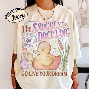Może przedstawiać: Kremowa koszulka z grafiką przedstawiającą kaczkę, trzciny i napis "The Snuggly Duckling Brewing Company". Projekt zawiera lampiony i frazę "Go Live Your Dream".