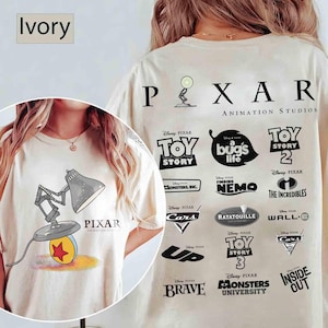 May include: Ivory-colored t-shirt featuring a Pixar lamp graphic and the word "PIXAR". The back of the shirt displays the Pixar Animation Studios logo and various movie titles, including "Toy Story" and "Finding Nemo".