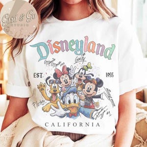 Puede incluir: Camiseta blanca con un dise&ntilde;o colorido de Disneyland con Mickey Mouse, Minnie Mouse, Goofy, Pluto y Donald Duck. El dise&ntilde;o incluye el texto "Disneyland California" y el a&ntilde;o "1955".