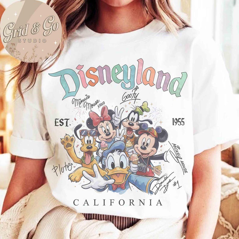 Puede incluir: Camiseta blanca con un dise&ntilde;o colorido de Disneyland con Mickey Mouse, Minnie Mouse, Goofy, Pluto y Donald Duck. El dise&ntilde;o incluye el texto "Disneyland California" y el a&ntilde;o "1955".
