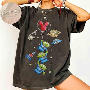 May include: Dark gray t-shirt featuring a colorful graphic of three green aliens in blue suits, holding onto a red Mickey Mouse balloon. The design includes a rocket, planets, stars, and a UFO.