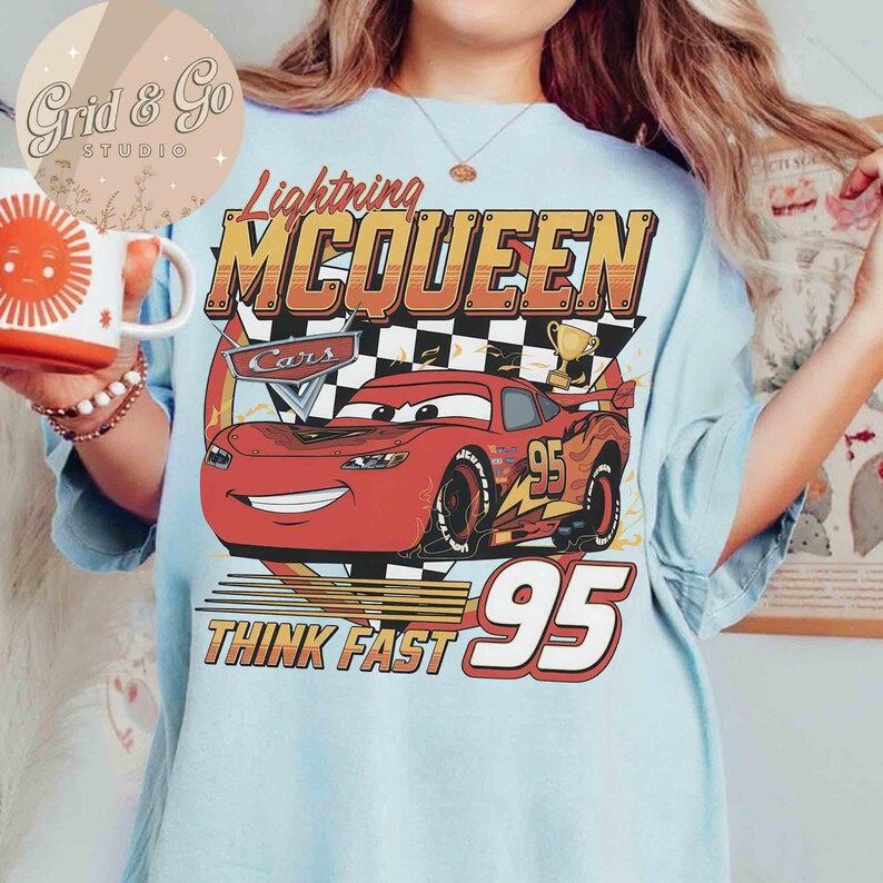 Pu&ograve; includere: T-shirt azzurra con un disegno di Saetta McQueen del film Cars. Il design include il testo "Lightning McQueen", "Cars", "95" e "Think Fast". La grafica &egrave; in rosso, giallo e nero.