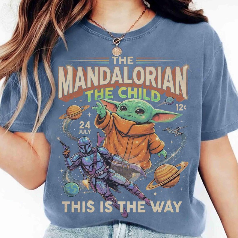 May include: Blue t-shirt with a graphic of The Mandalorian and Grogu from Star Wars. The design includes planets, a spaceship, and the text "THE MANDALORIAN THE CHILD" and "THIS IS THE WAY".