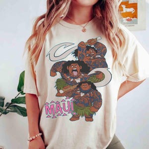 May include: Cream-colored t-shirt featuring a graphic of the character Maui from the animated film Moana. The design includes three Maui figures with tattoos, a fish hook, and the word "MAUI" in pink letters.