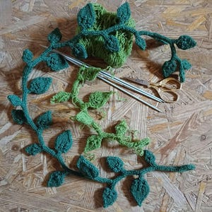 Peut inclure: Divers éléments de crochet faits à la main, dont des vignes de feuilles vertes, de la laine, des aiguilles à tricoter et des ciseaux dorés. Les articles sont disposés sur une surface en bois, mettant en valeur les fournitures et les pièces finies.