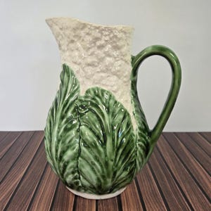 May include: A ceramic pitcher with a textured, off-white upper section and a green handle. The lower portion features a detailed, green cabbage leaf design. The pitcher is set on a wooden surface.