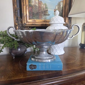 May include: A silver-colored, scalloped-edge bowl with two handles, resting on a pedestal base. The bowl sits atop a blue book. A white decorative jar and a framed painting of a canal scene are in the background.