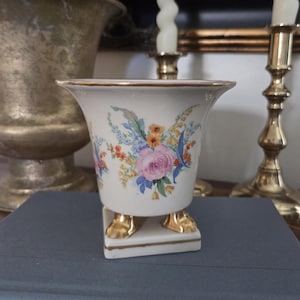 May include: A white porcelain vase with a floral design featuring pink roses and blue accents. The vase has gold trim and rests on gold-colored feet. The vase is sitting on a blue book.