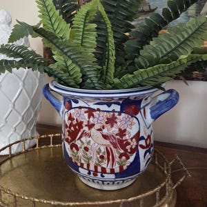 May include: A decorative ceramic planter with a bird and floral design in red, white, and blue. The planter has two blue handles and is filled with green fern plants. The planter sits on a gold-colored tray.