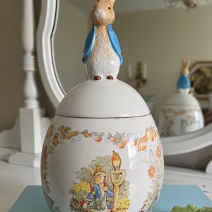 May include: A ceramic jar with a removable lid featuring a sculpted Peter Rabbit figure wearing a blue jacket. The jar is white and decorated with a colorful illustration of a rabbit in a garden with a shovel and bird, surrounded by orange flowers.