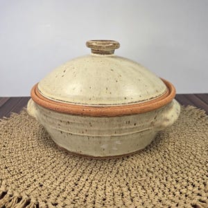 May include: A beige ceramic lidded casserole dish with a brown rim and handle. The lid has a small, round handle. The dish is sitting on a woven placemat.