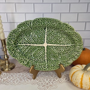 May include: An oval ceramic serving platter shaped like a green cabbage leaf with a glossy finish and a prominent white central vein structure. The platter features a textured surface that mimics the natural appearance of layered cabbage leaves.