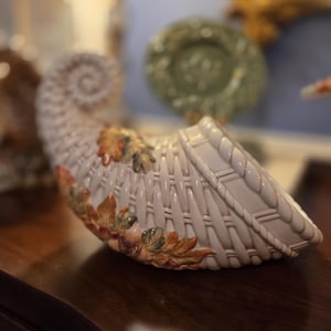 May include: A ceramic cornucopia in a cream color with a woven basket design. The cornucopia is decorated with autumn leaves in shades of orange, brown, and green. The horn of plenty is displayed on a dark wooden surface.