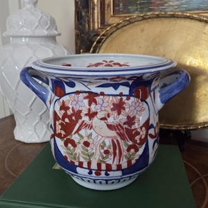 May include: A ceramic planter with a blue rim and handles, featuring a detailed design of a bird and flowers in red, white, and blue. The planter sits on a green book with gold lettering. A white decorative jar is in the background.