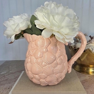 May include: A light pink ceramic pitcher featuring a textured, embossed surface covered in various seashell patterns. The pitcher has a curved handle and is filled with large, white artificial peony flowers with green leaves.